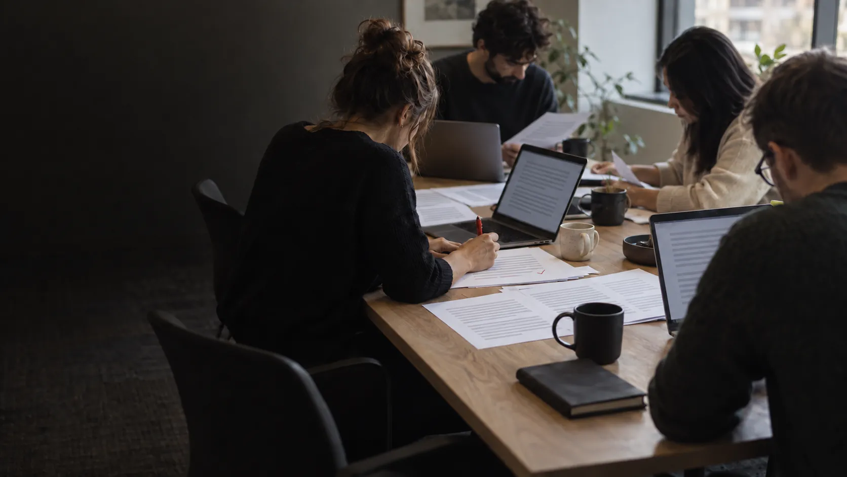 Four people at one table, each working through printed pages and their own laptop.