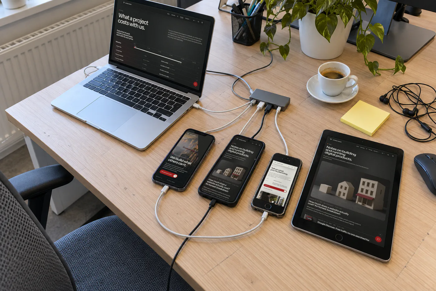 The corner of a desk with three different phones and a tablet plugged into one hub beside a laptop, each showing a page of our website, next to an espresso and a blank sticky-note pad.