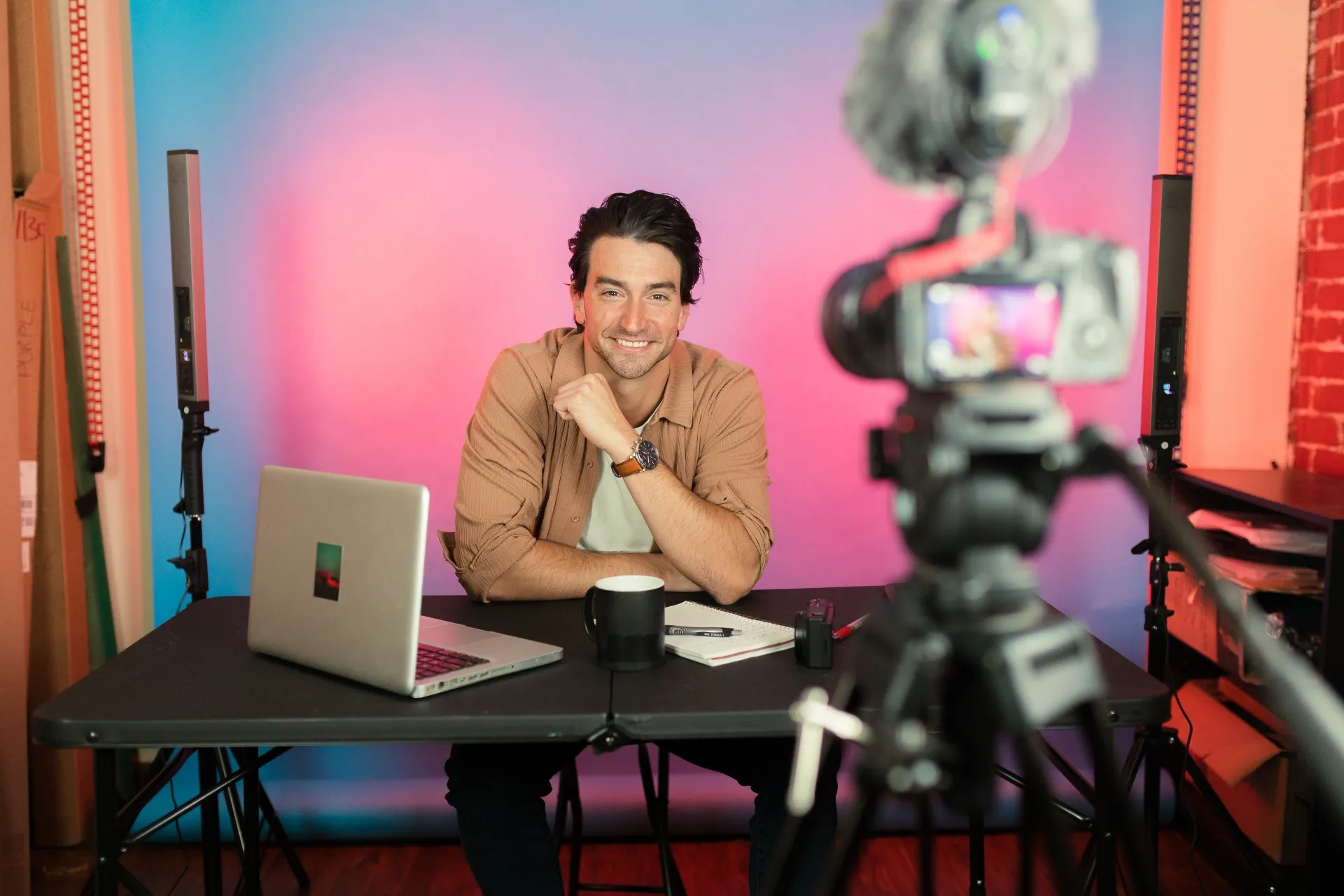 Content creator recording video at desk with laptop, DSLR on tripod, pink‑blue studio lights behind.
